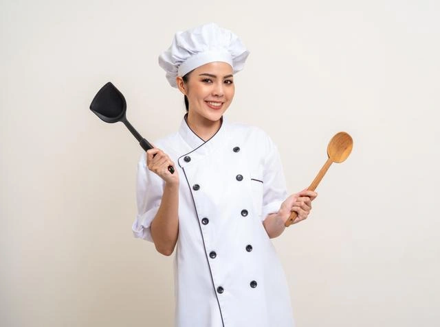A professional chef holds a cooking spoon in her left hand and a spatula in her right.