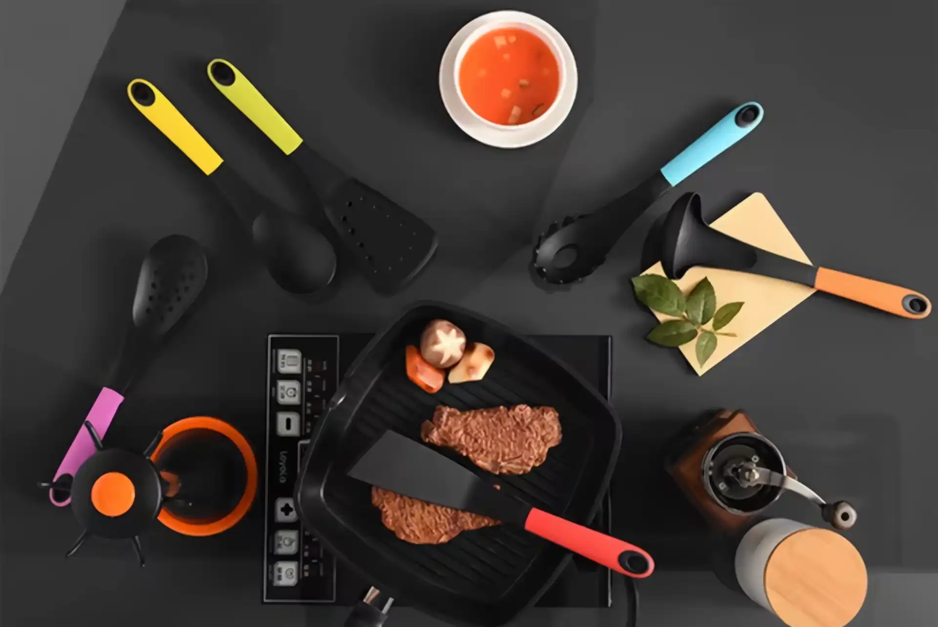 Cookware and black nylon cooking utensils with colors handle sit on a light black countertop