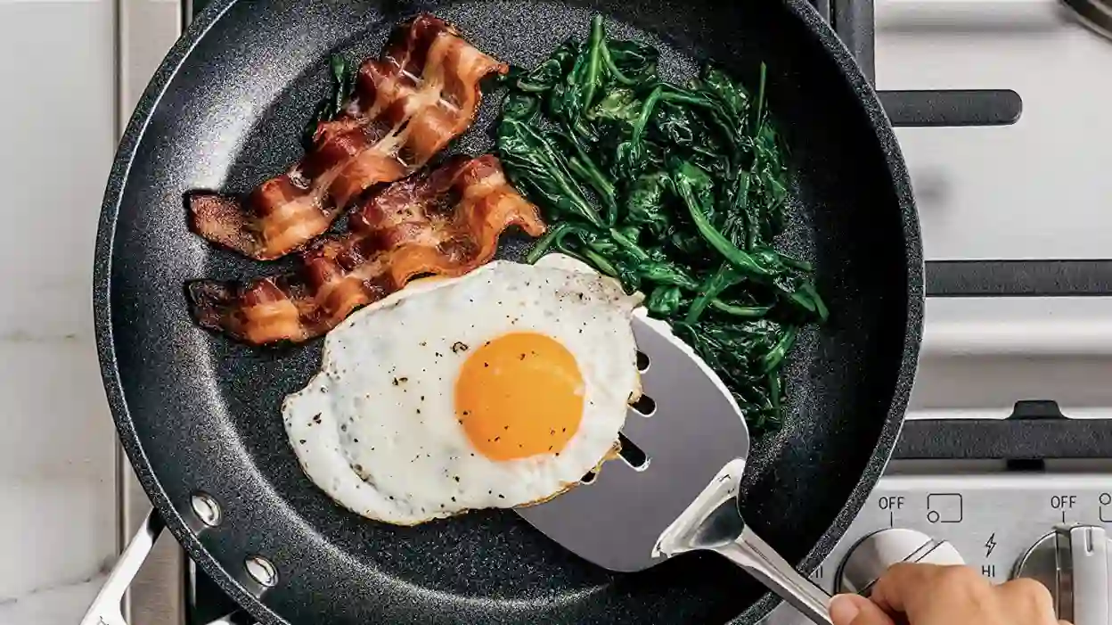 One hand was frying eggs and bacon in a nonstick pan with a metal spatula